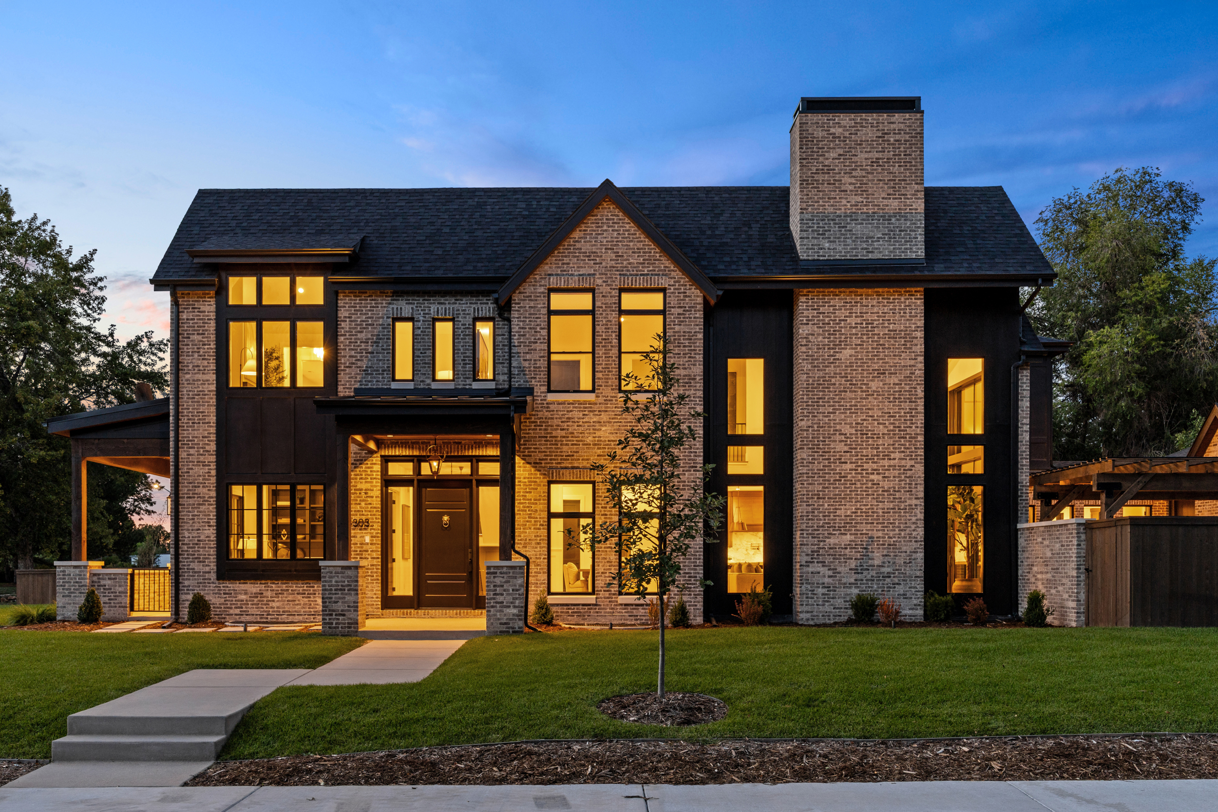 Modern two-story brick and dark wood home at dusk with warm interior lighting and a manicured green lawn.